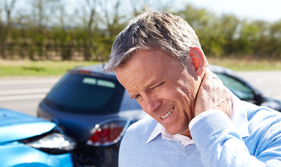 A man holds a hand to the back of his neck while wincing in pain while two cars are collided behind him