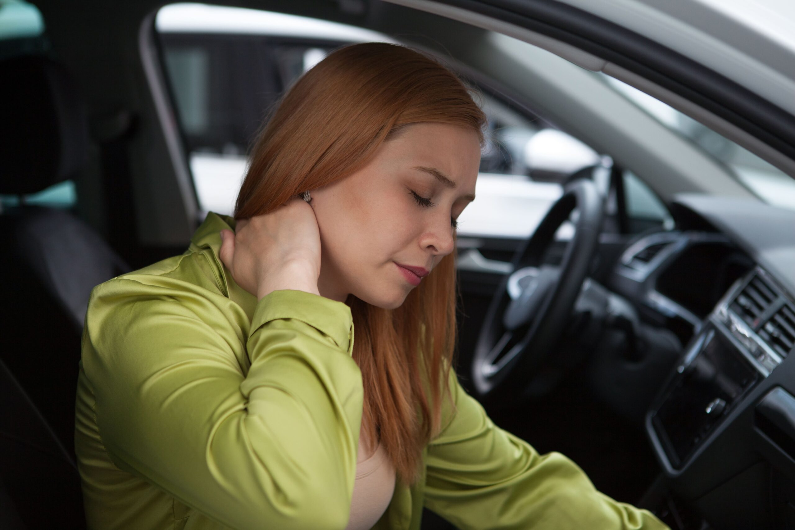 A woman in a green top sits in a car while holding the back of her neck with her right hand, wincing in pain.