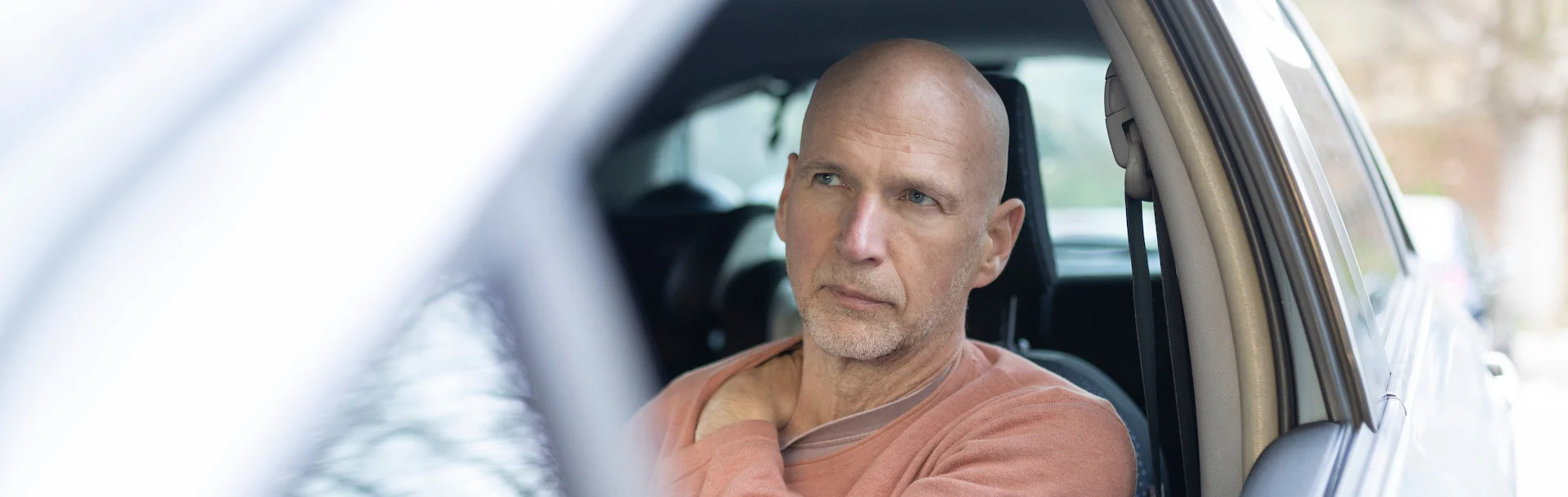 A man in a salmon-colored sweat shirt grips his shoulder while sitting in the car.
